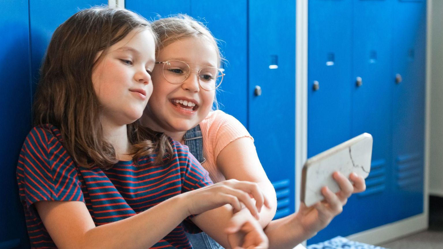 Two primary school-aged girls sitting with their backs to blue lockers, one is holding out a smartphone which they are both looking at with cautiously smiling faces