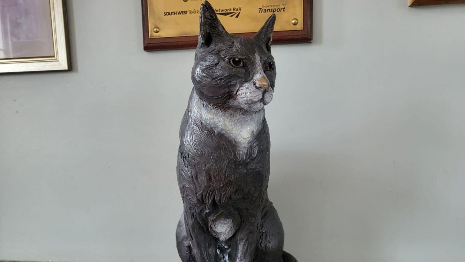 A close-up shot of a life-size statue of a black and white cat against a grey wall, with the bottom edges of plaques and pictures just visible above it