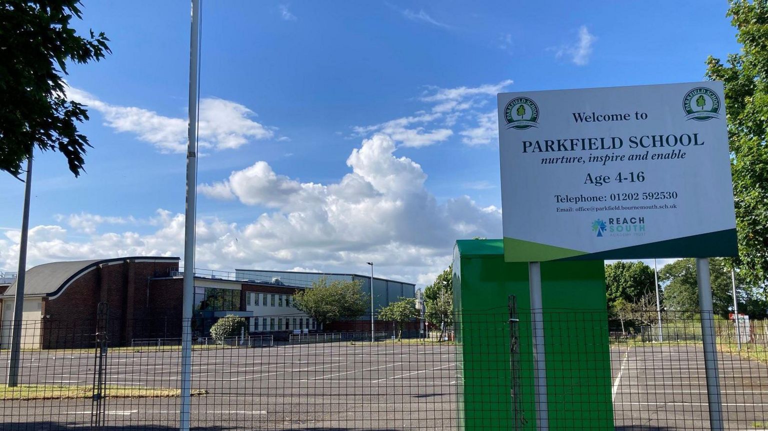 A sign saying 'welcome to Parkfield School' with an empty car park and a building in the background 