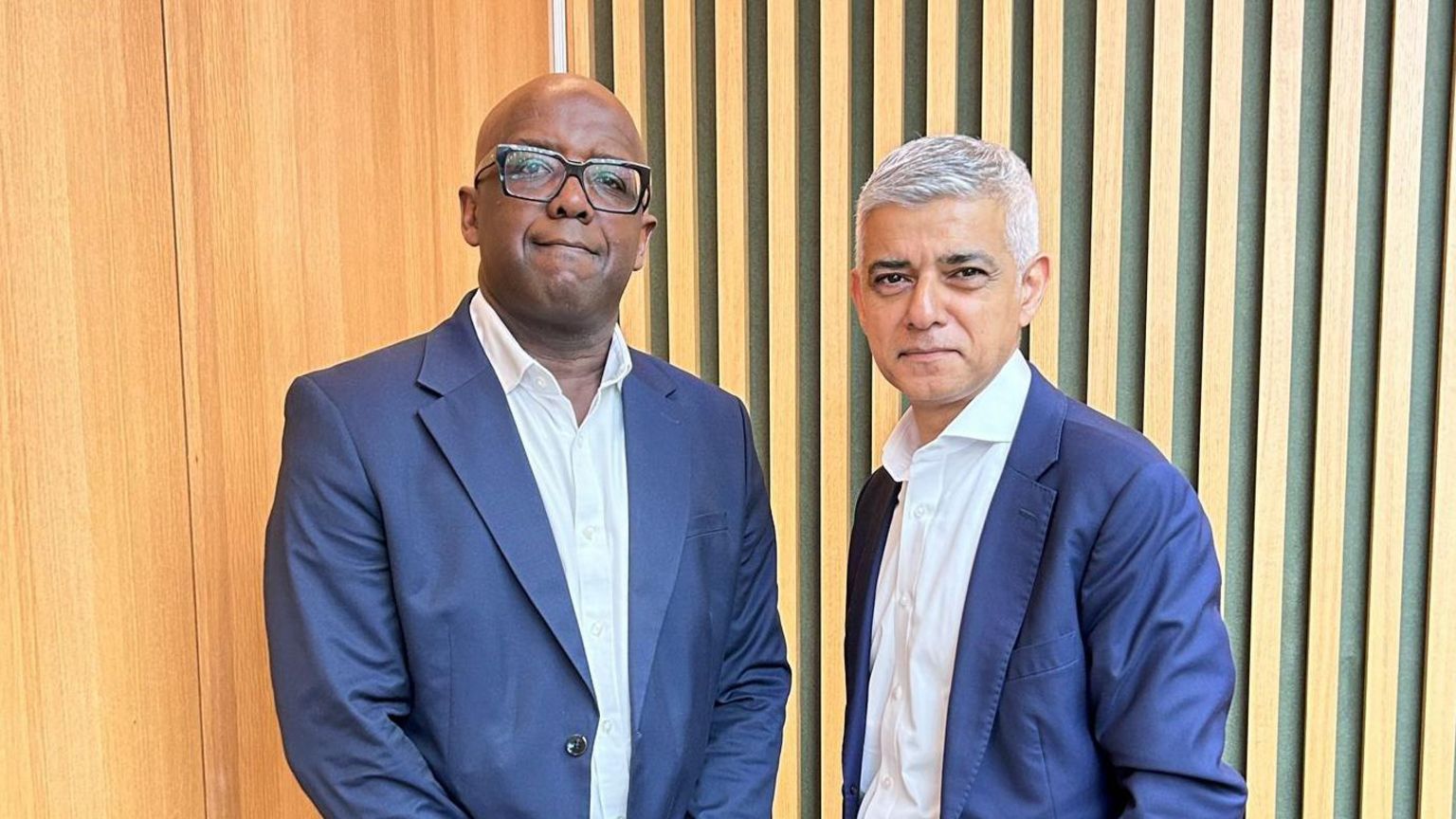 Trevor Sterling standing next to Sadiq Khan. They're both standing against a wood panelled wall, wearing blue suit jackets and white shirts. Mr Sterling is wearing glasses.