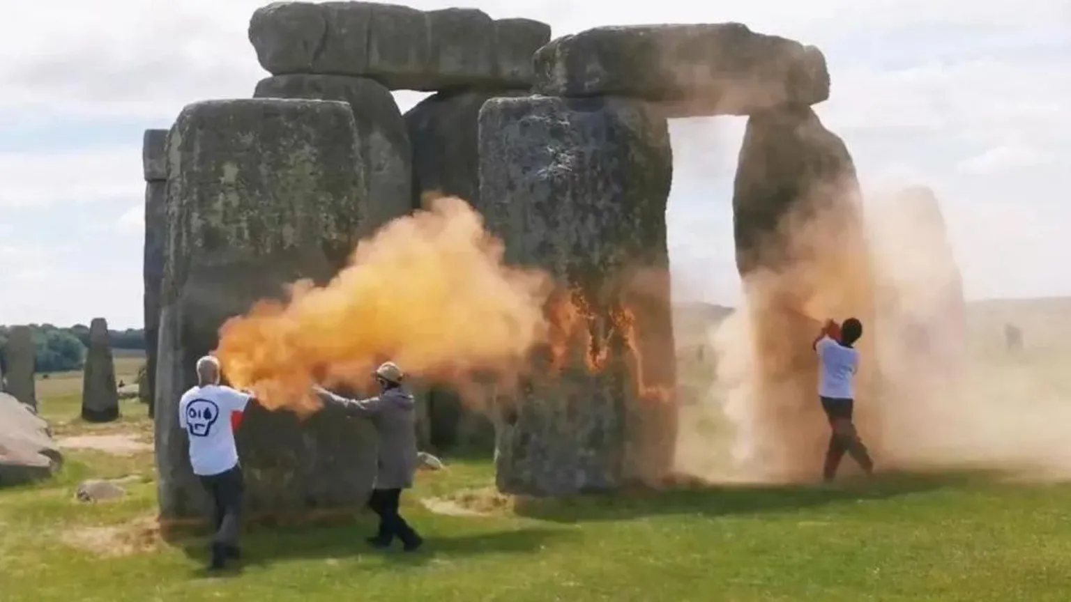 The image shows Stonehenge's ancient stone trilithons covered in bright orange powder paint. Two people in white shirts are visible near the monument, with one appearing to spray or apply the paint while orange clouds billow around the stones. The prehistoric structure stands on green grass under a cloudy sky.