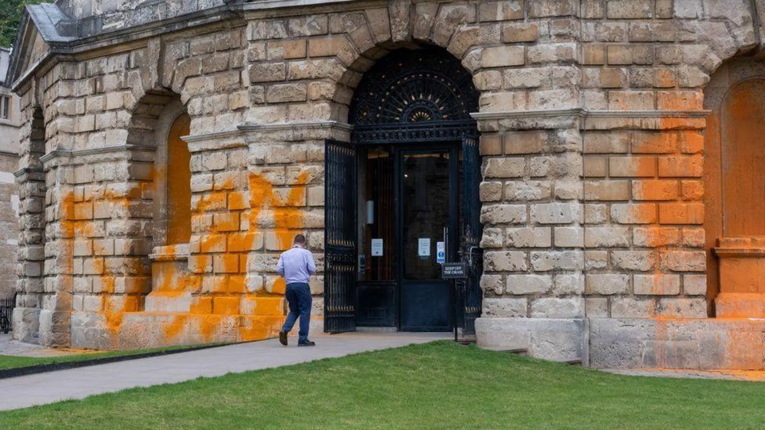 The image shows a historic stone building with classical architecture featuring arched openings and columns. Orange paint has been sprayed across the stonework in large patches. A person in light clothing is walking near the entrance, which has ornate black doors with decorative metalwork above.