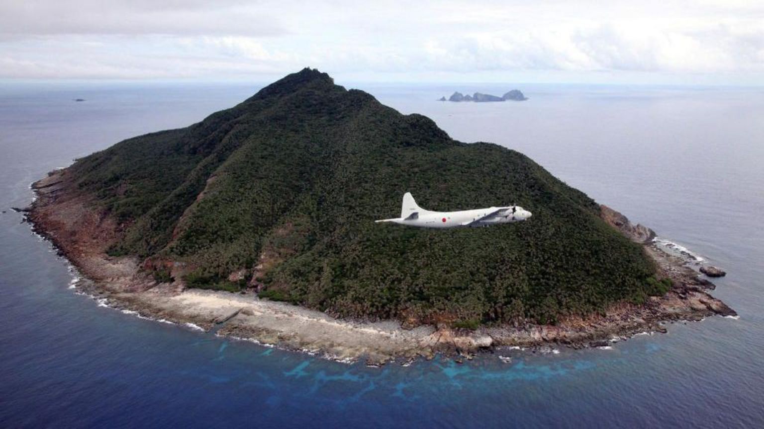 一架白色飞机在空中倾斜飞行,掠过郁郁蔥蔥的丘陵岛屿,该岛屿属于尖阁/钓鱼岛群岛的一部分。