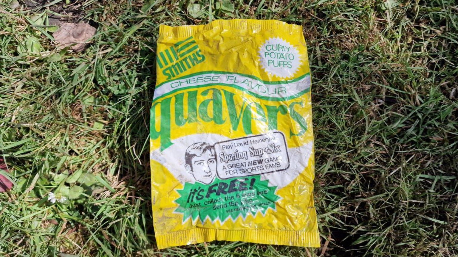 An old quavers crisp packet in yellow, green and white colours, on a patch of green grass