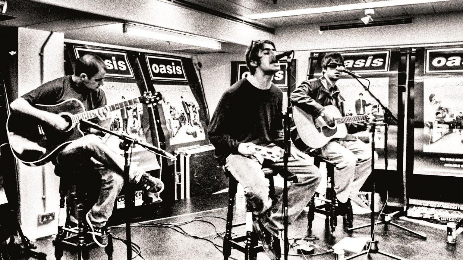 Black and white photo of Bonehead (left), Liam Gallagher (centre) and Noel Gallagher (right) sitting on high stools with microphones in front of them. Bonehead and Noel are playing guitars and Liam is holding a tambourine as he sings into the microphone. There are large Definitely Maybe posters behind them and to the right