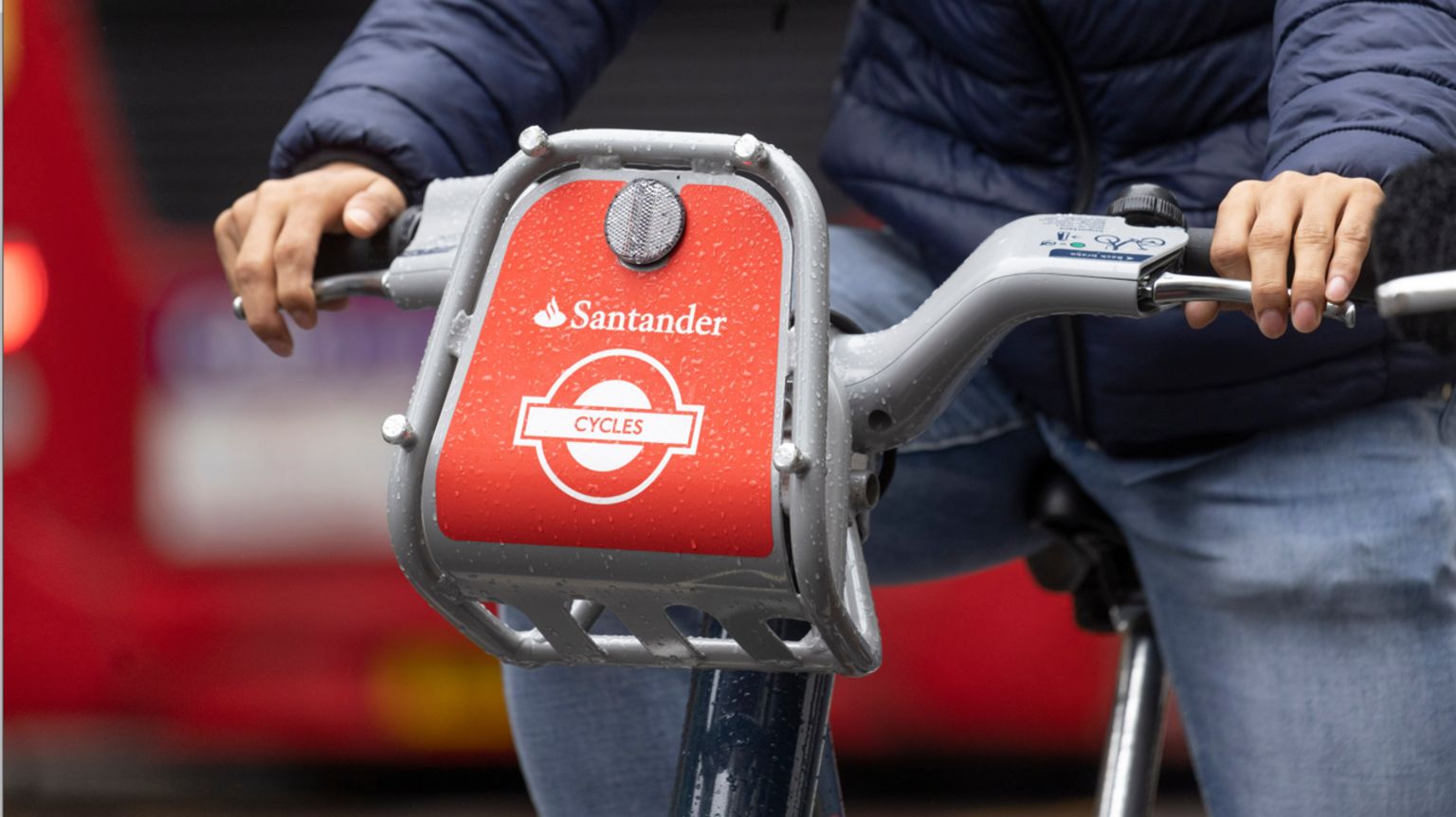 Red Santander branded e-bikes ridden by someone wearing jeans and a blue puffer jacket