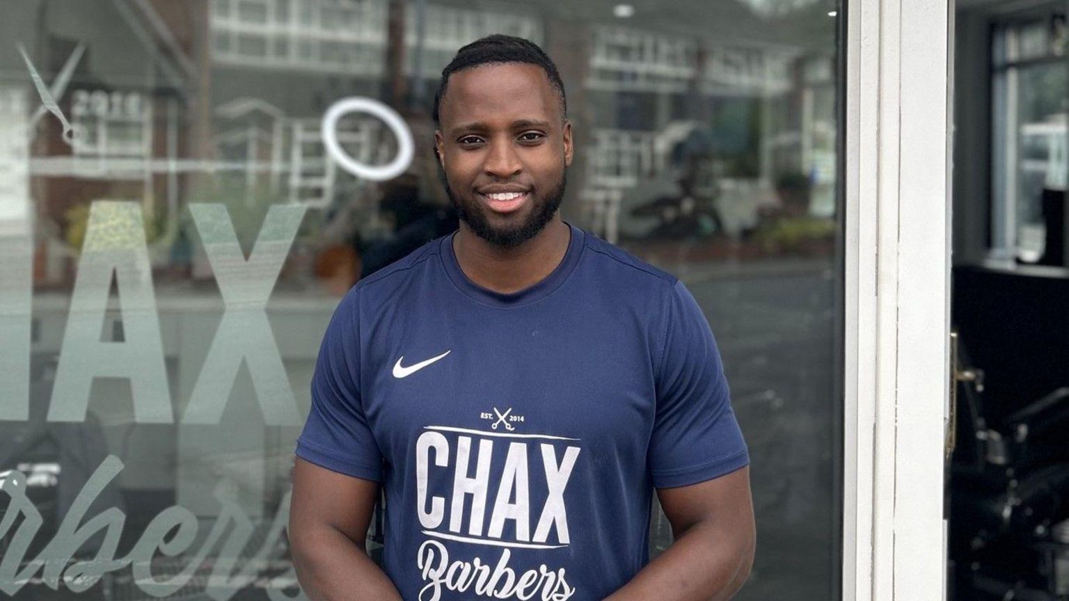 Leeroy Chax standing in a blue T-shirt outside his shop, both branded with the name of the shop, Chax Barbers.