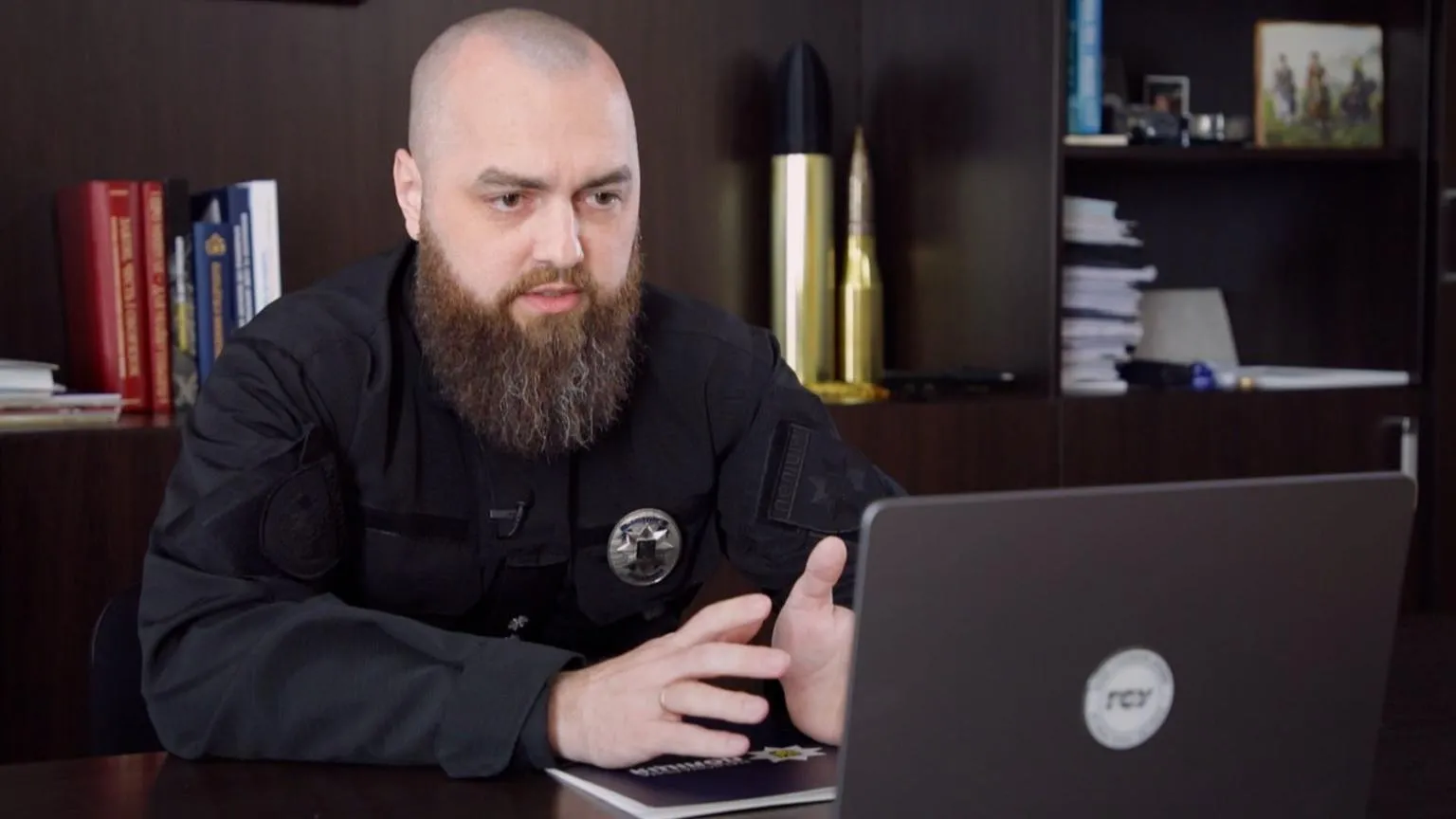 Vitaliy Sova, a man with a shaved head and a long brown beard, speaking on a video call in his laptop while he is filmed from a different angle in the office, which has black bookshelves behind him.