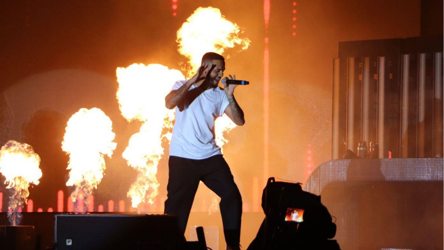 Aston Merrygold from JLS standing on stage wearing a white t-shirt and dark trousers. He is holding a microphone in one hand and gesturing with the other, while fire canons go off in the background