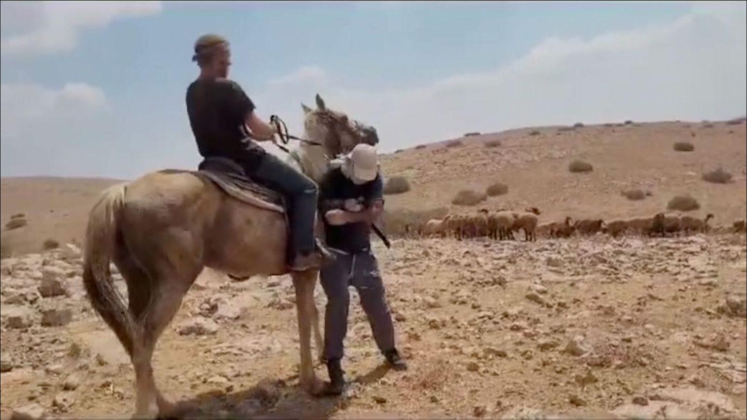 Gil appears to be standing his ground as a settler on horseback rides into him. They are in the dusty landscape of the Jordan Valley, and a flock of sheep can be seen in the middle distance.