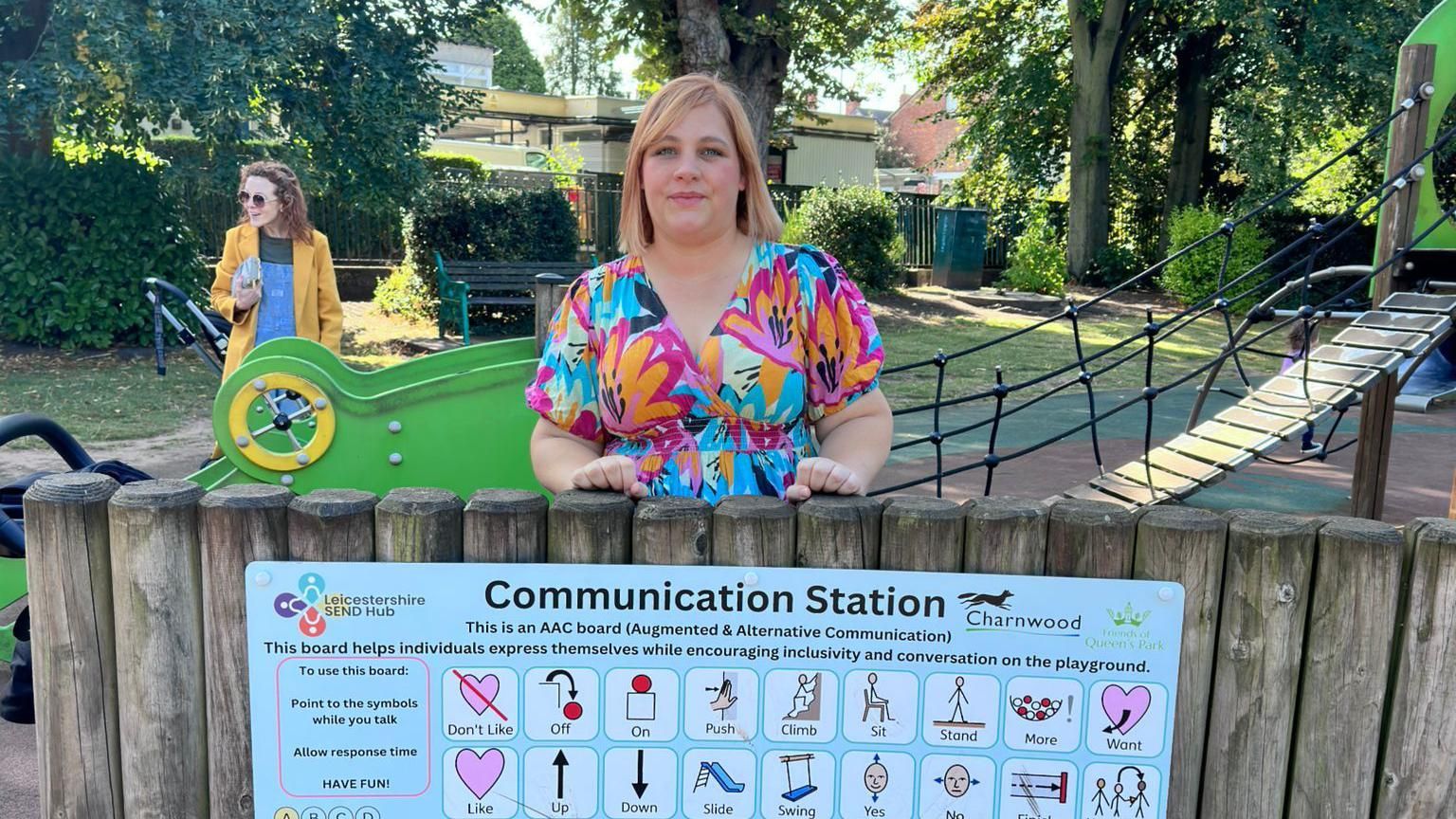 Megan Stewart standing behind the communication board, which is on a fence. The board has symbols on including push, climb, and stand, as well as the alphabet 