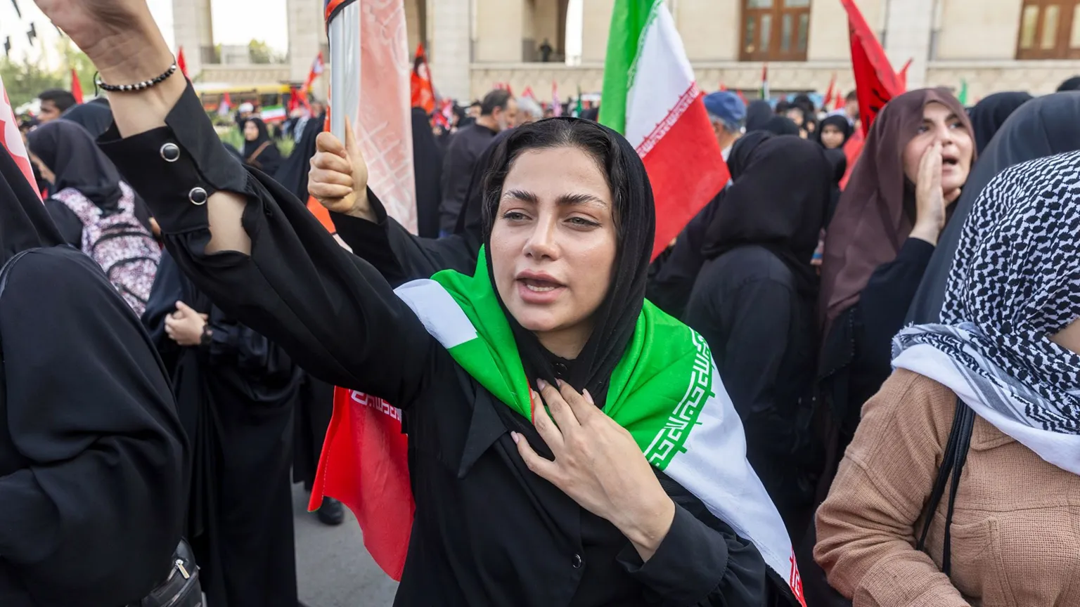 A young Iranian woman in Tehran, with an Iranian flag draped over her shoulders, raises her hand in a crowd of other people shouting and waving flags
