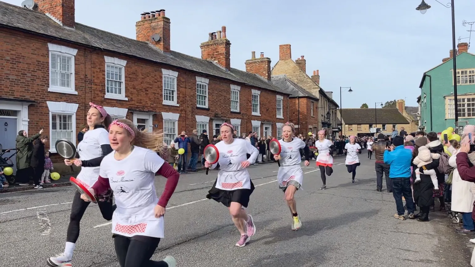 A group of women in aprons and themed outfits run down a street while holding frying pans with pancakes, taking part in The Olney Pancake Race. Spectators line the roadside, watching and taking photos in a residential neighborhood.