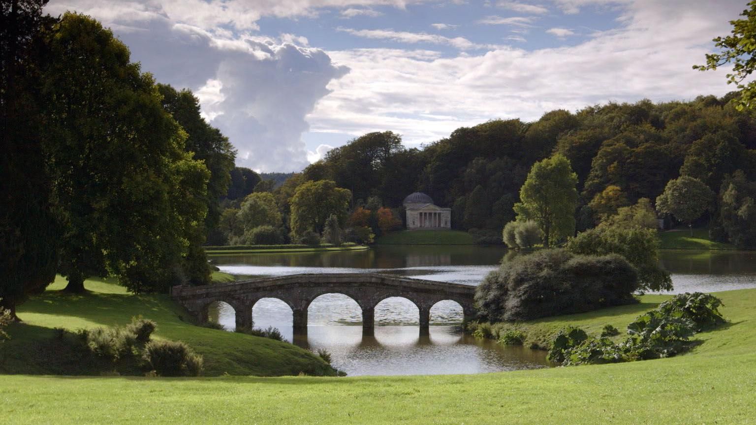 A picture of the lake on the Stourhead estate, which has a classically-inspired building in the background and a small arched bridge in the foreground. 
