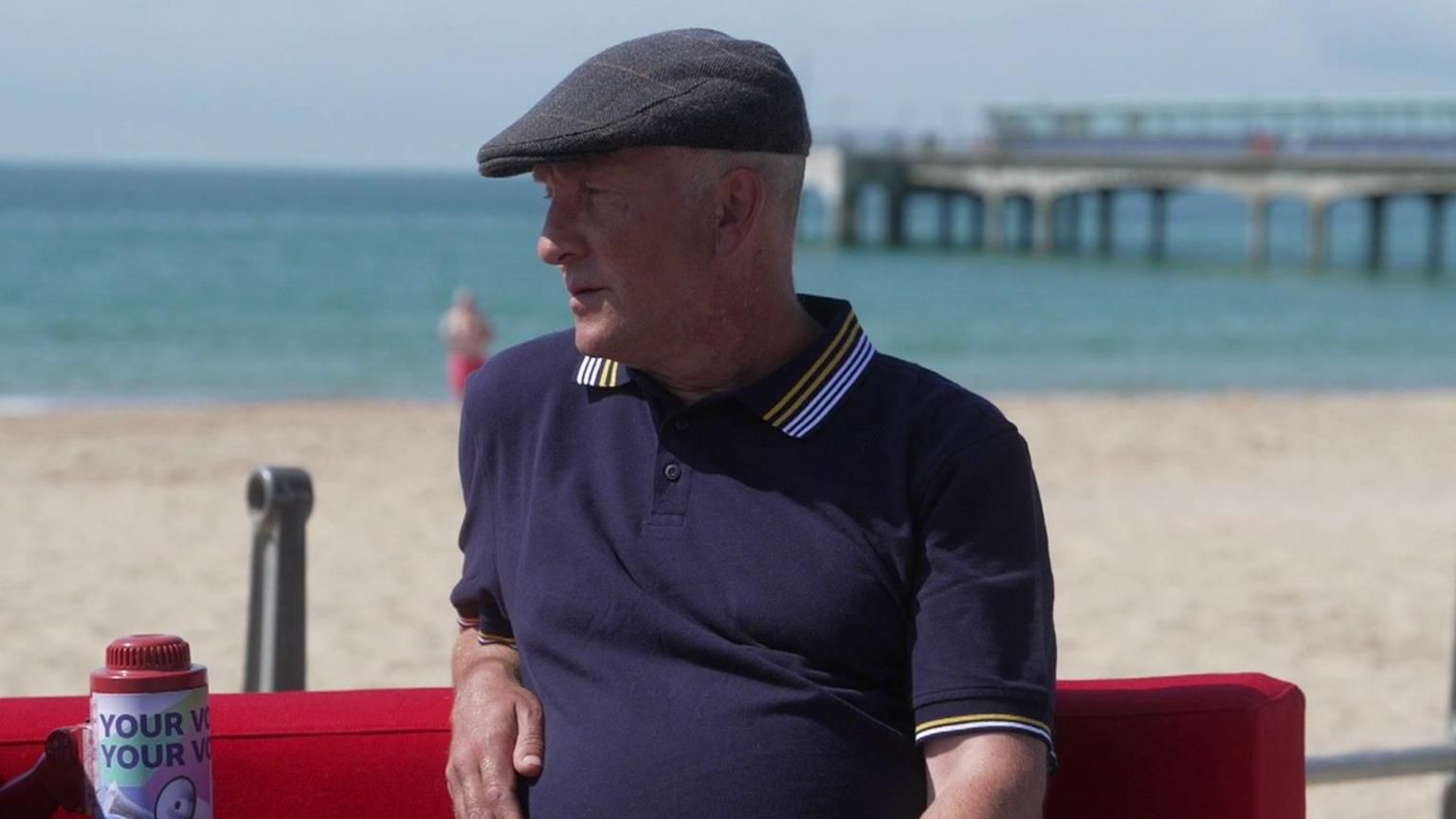 Andrew wears a dark blue polo shirt with a grey, checked hat and is sitting on a red sofa with Bournemouth beach behind him