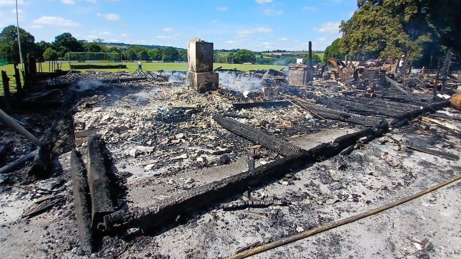 Charred wooden beams are lying on a bed of smoking ashes and rubble with the sunny green cricket pitch visible beyond