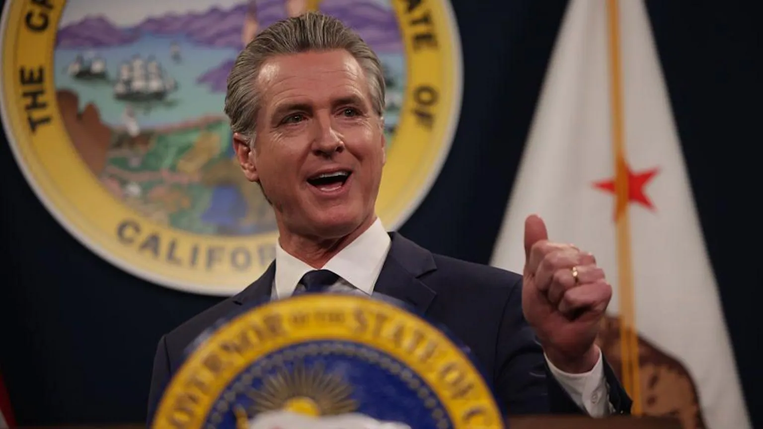 San Francisco Chronicle via Getty Images California Governor Gavin Newsom speaks at a podium bearing the California state seal. The California flag, which features a grizzly bear and a red star, is vi