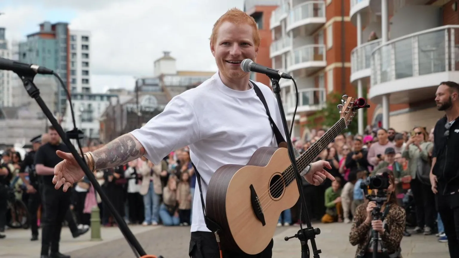Shaun Whitmore/BBC Ed Sheeran performing at Ipswich Waterfront. He is performing to crowds of hundreds of people with a microphone and guitar. He is wearing a white T-shirt and is looking at the camera and smiling.