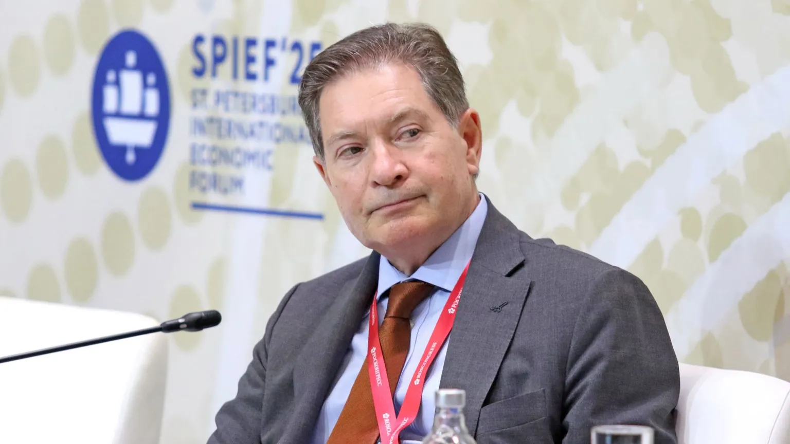 Getty Images Man with brown and greying hair, wearing a grey suit with blue short and brown tie. A red lanyard is around his neck.