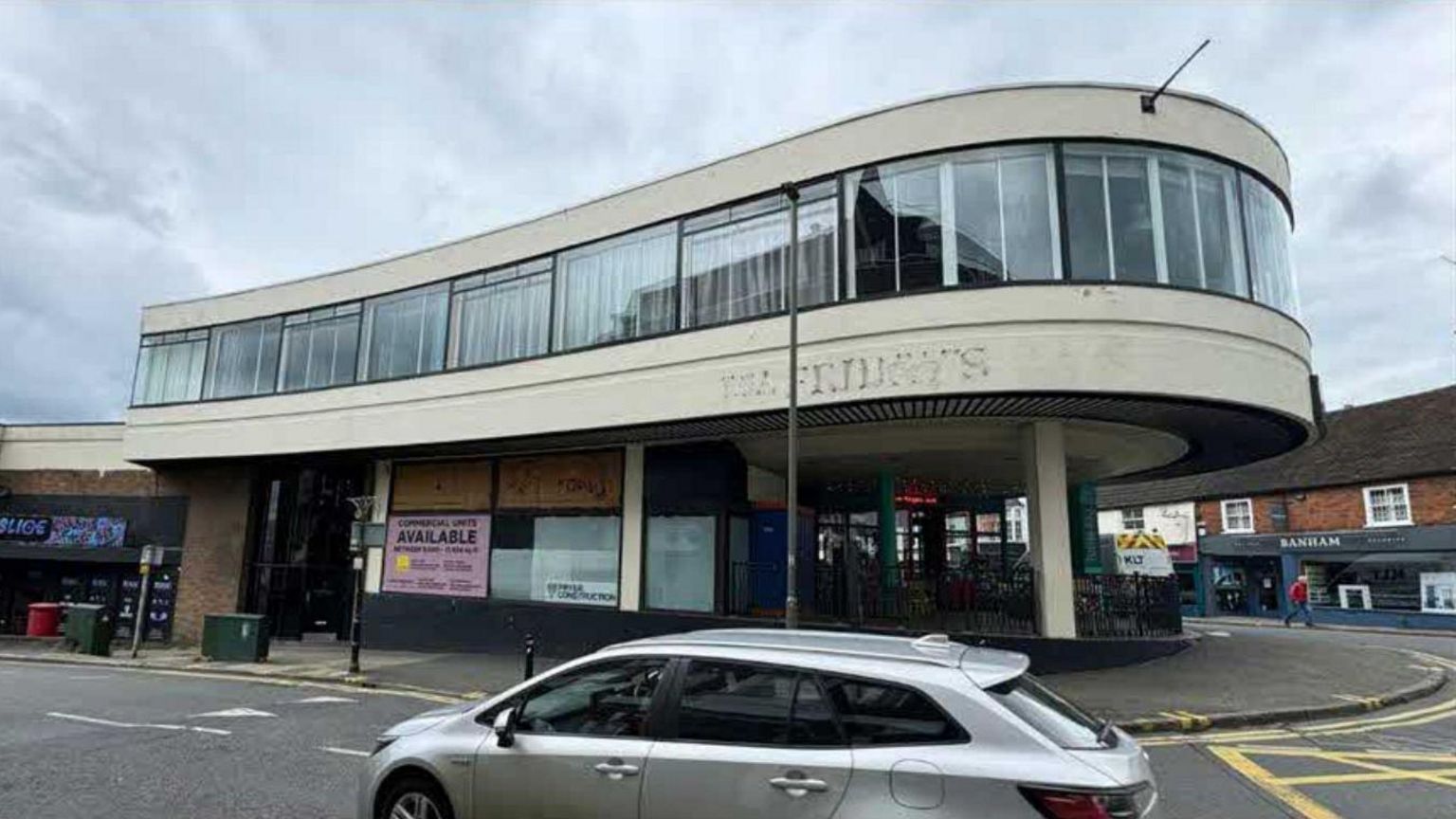 A concrete grey curved building, with glass around the second story. A silver car is passing by 