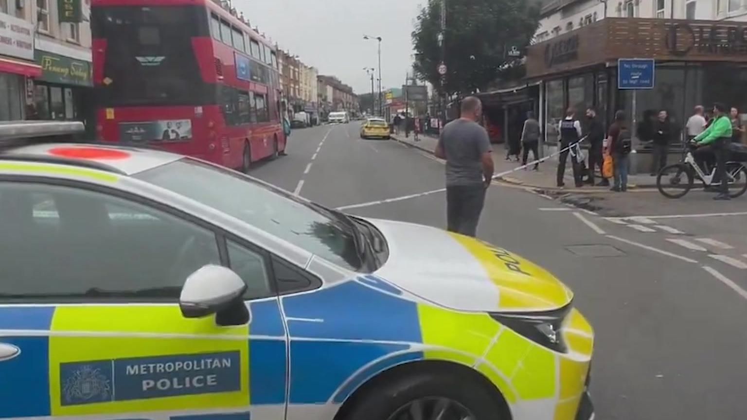 Police car and tape and onlookers on Turnpike Lane. In the foreground there is a police car and behind it some police tape cordoning off a section of road near a junction.