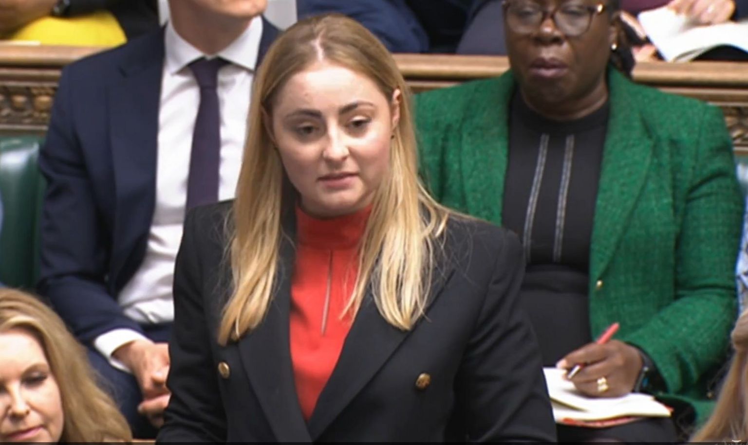 Gen Kitchen, with long blond hair, wearing red top and black jacket standing in the House of Commons