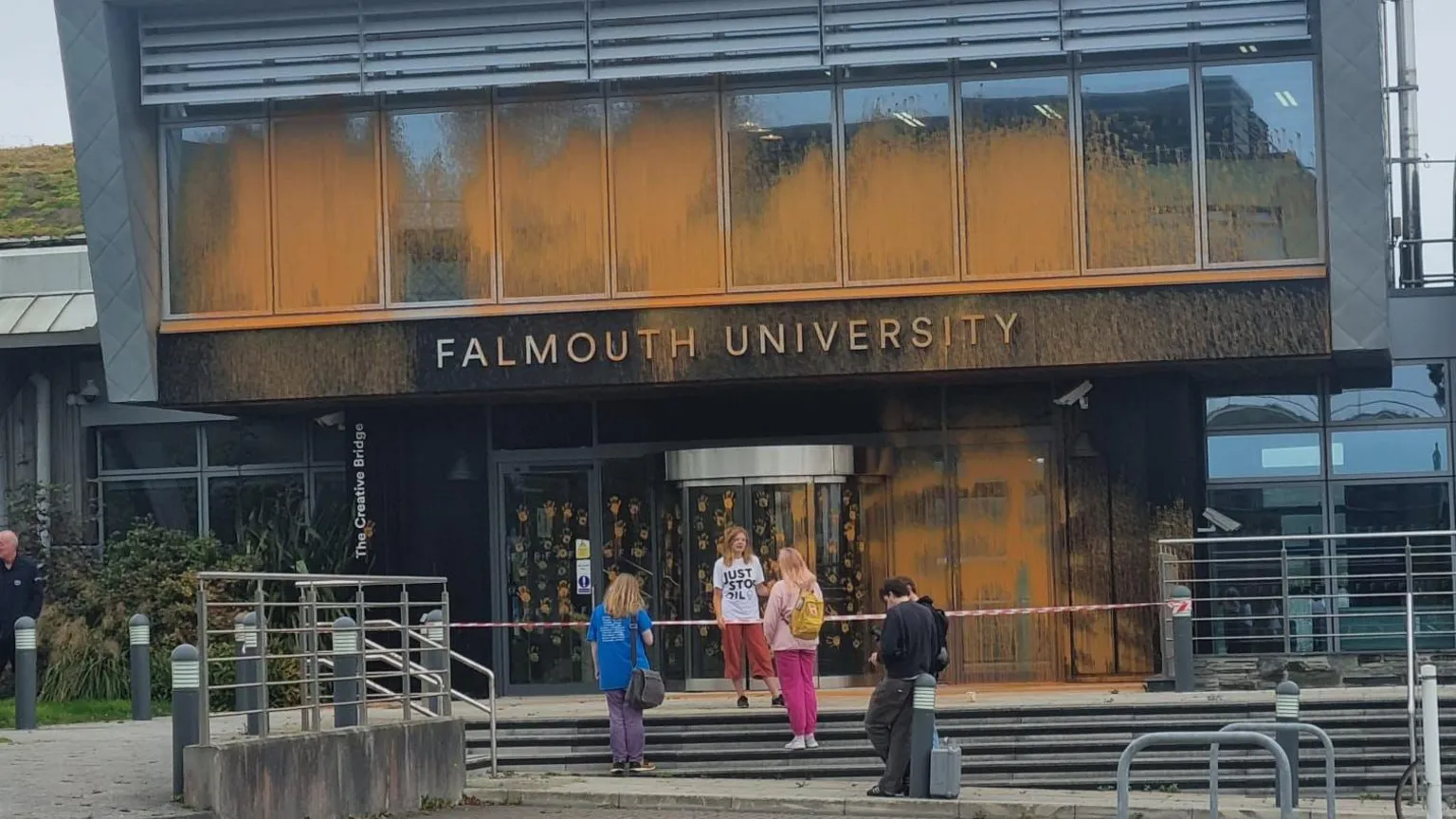The image shows Falmouth University's modern glass-fronted building with orange paint splattered across its windows and entrance area. Several people, including students in colorful clothing, are gathered near the entrance steps. Red and white barrier tape is visible, cordoning off the affected area after the paint incident.
