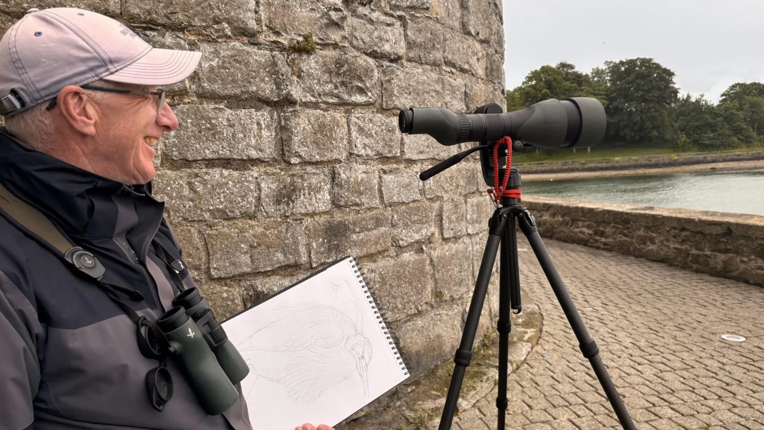 Richard Partis Richard Partis looks out at water smiling. He holds a sketchbook and stands behind his camera lense. He wears a grey jacket, black binoculars and a grey cap.