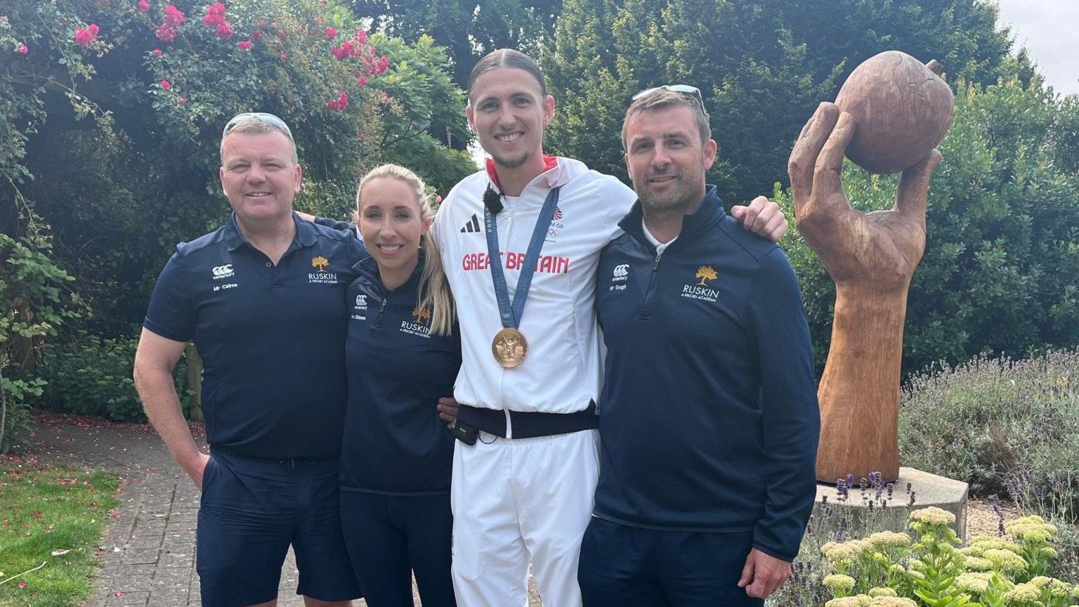 Lewis Davey poses for a photo with his former PE teachers. Adam Cairns has short, light brown hair and has sun glasses on his head. He is standing with his right hand in his pocket and his left hand around the waist of Emma Gibbons. She has blonde hair in a pony tail and is smiling at the camera. Lewis is wearing his white Great Britain tracksuit with his medal around his neck and is stood next to Rob Gough, who has a long-sleeve tracksuit top on and glasses on his head. 