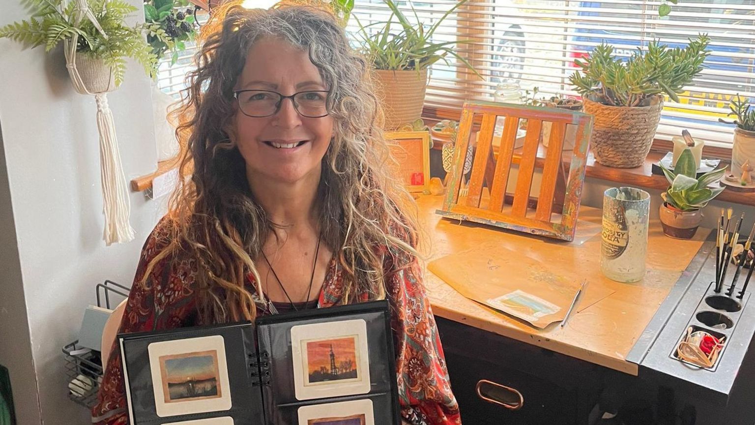 Caroline West - who has long, wavy grey and brown hair smiles at the camera - is holding a binder which is open to a page of painted teabags. Behind her is a desk with paintbrushes on