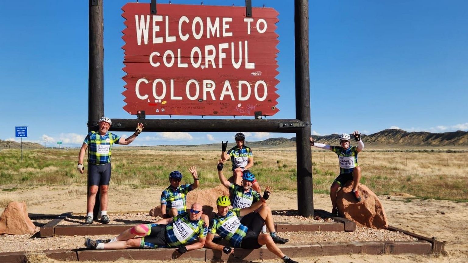 A group of men in brightly coloured cycling outfits posing in front of a sign that reads 'Welcome to Colorado'