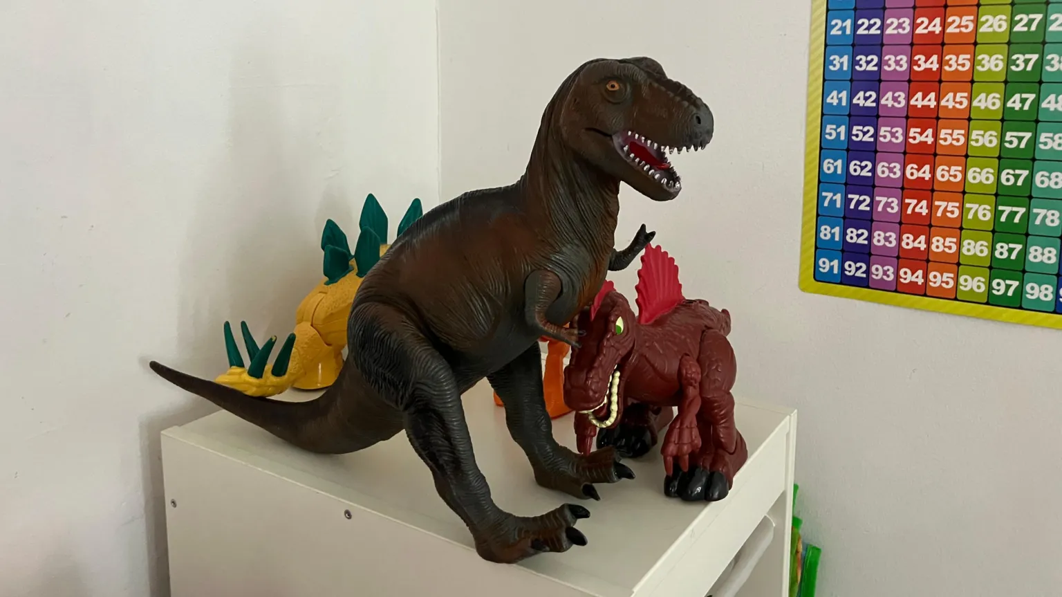 Vikki Irwin/BBC Three toy dinosaurs perched on the top of a white cabinet. On the right of the image is a colourful counting chart.