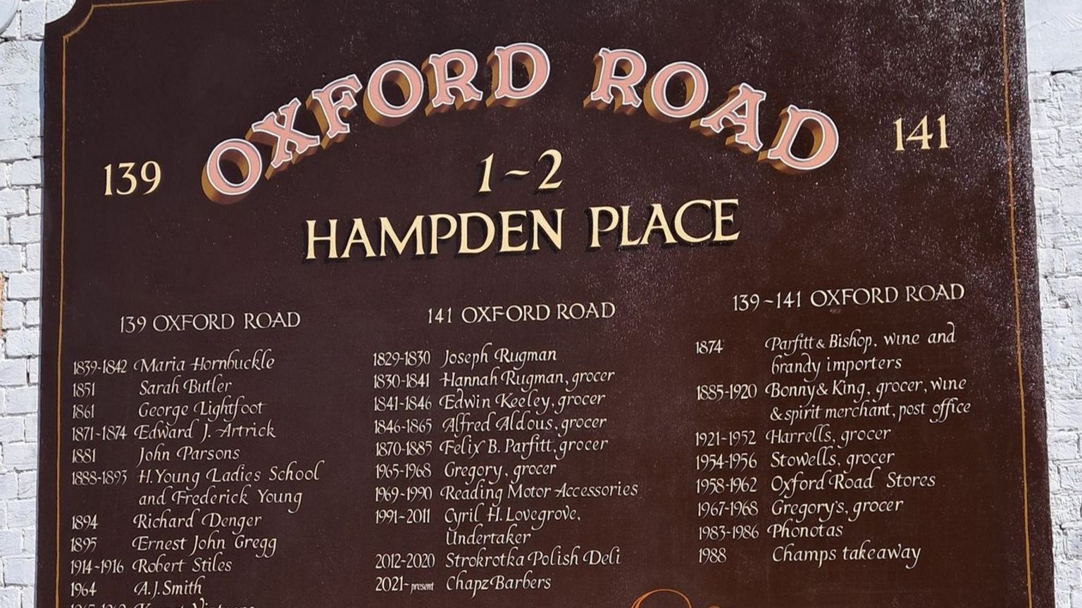 A dark red painted sign on a brick wall which has the dates and names of previous shops that have traded on Oxford Road.