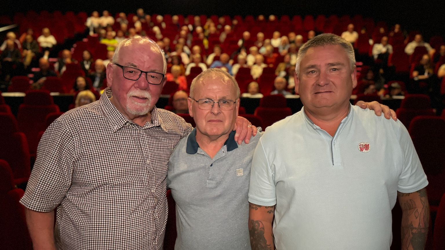 Three grey-haired men wearing short-sleeved shirts stand in front of an auditorium 