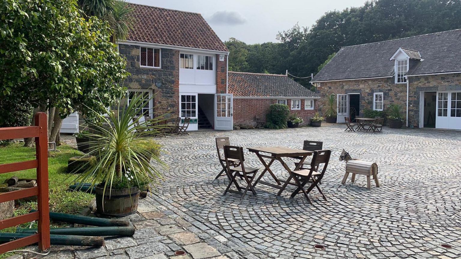 The courtyard of the folk museum in Saumarez Park. A cobbled courtyard, with tables and chairs. Small agricultural buildings and plants surround it.