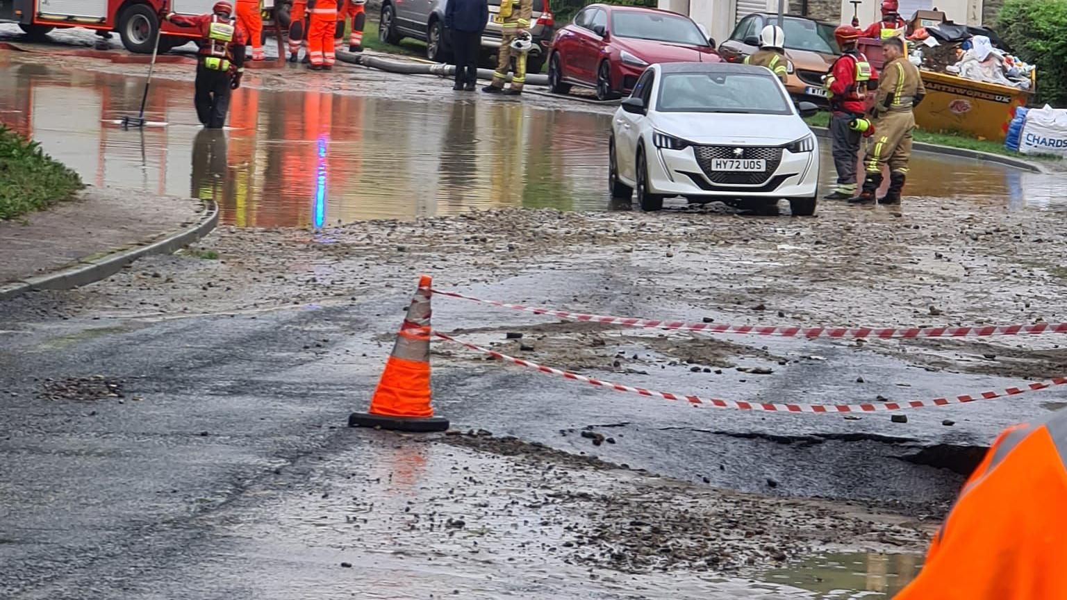 A road which has a large dent in it, which has been corned off by red and white striped tape and a traffic cone. A white car is driving on the road by the flood water and two fire fighters are standing next to the car.