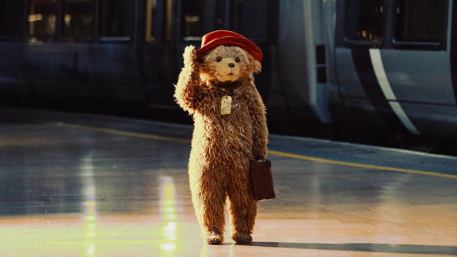 PA Small bear with red hat and suitcase stands alone on a train platform beside a parked train