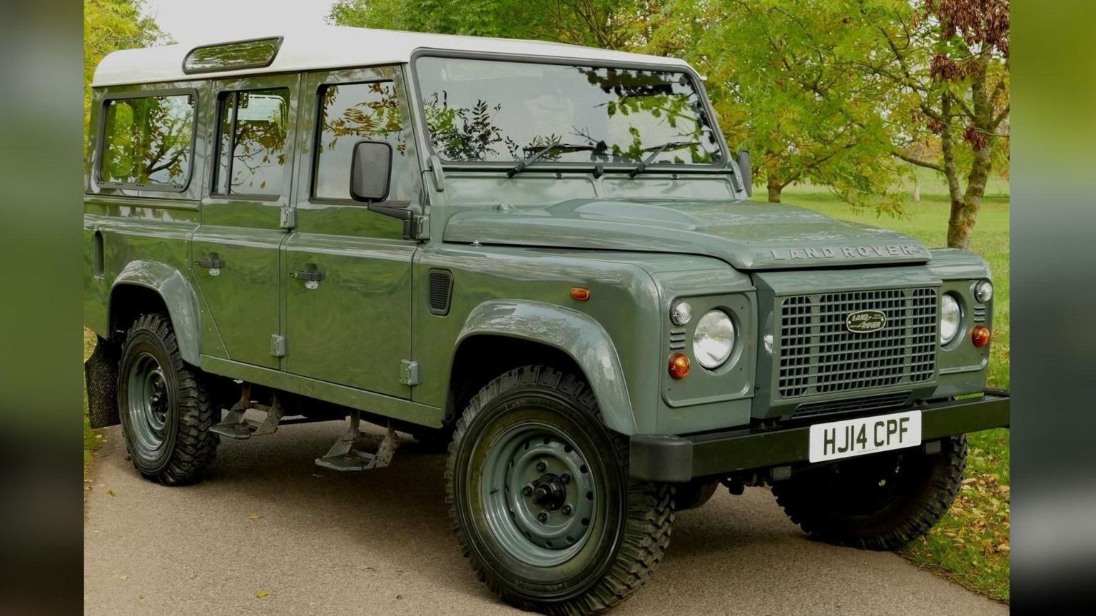 A green Land Rover Defender parked on a path with trees and grass visible in the background