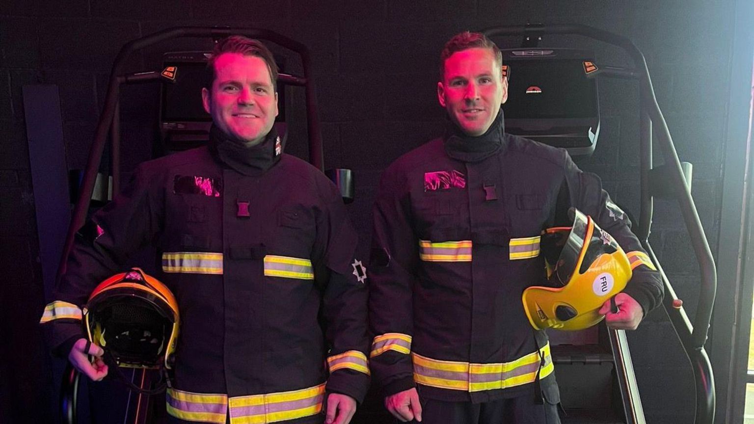 Sam Hayward and Jonathan Freeman smiling at the camera wearing full fire gear in front of two stair climbing machines