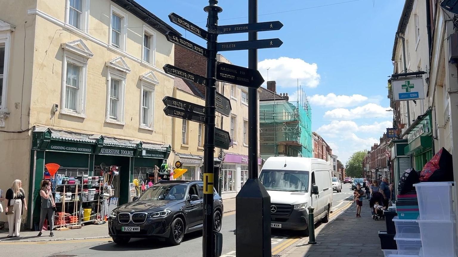 A signpost in the centre of Atherstone with a van and a car driving behind it in front of several shops