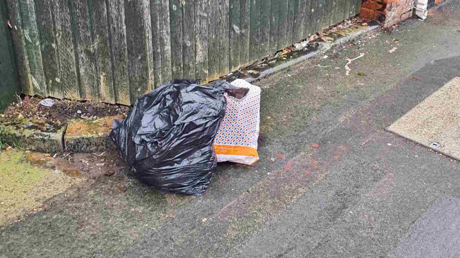 A black bin bag and a blue, white and orange carrier bag have been left on a pavement next to a fence.