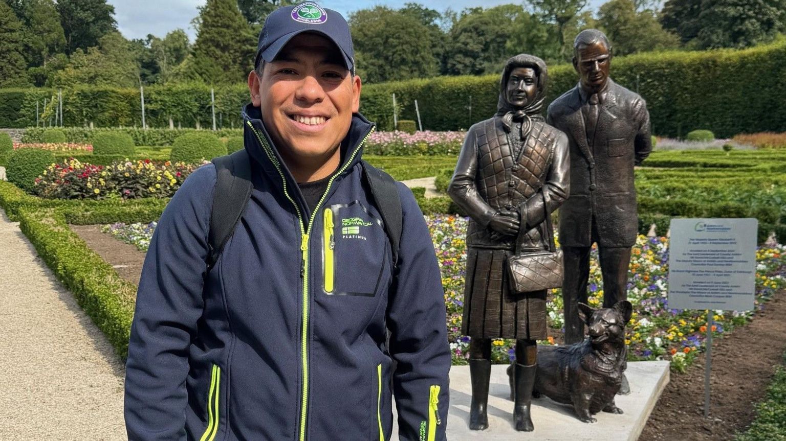 Alejandro is standing smiling in front of the statue. 

He is wearing a navy Wimbledon hat, navy waterproof jacket with florescent yellow zips.  