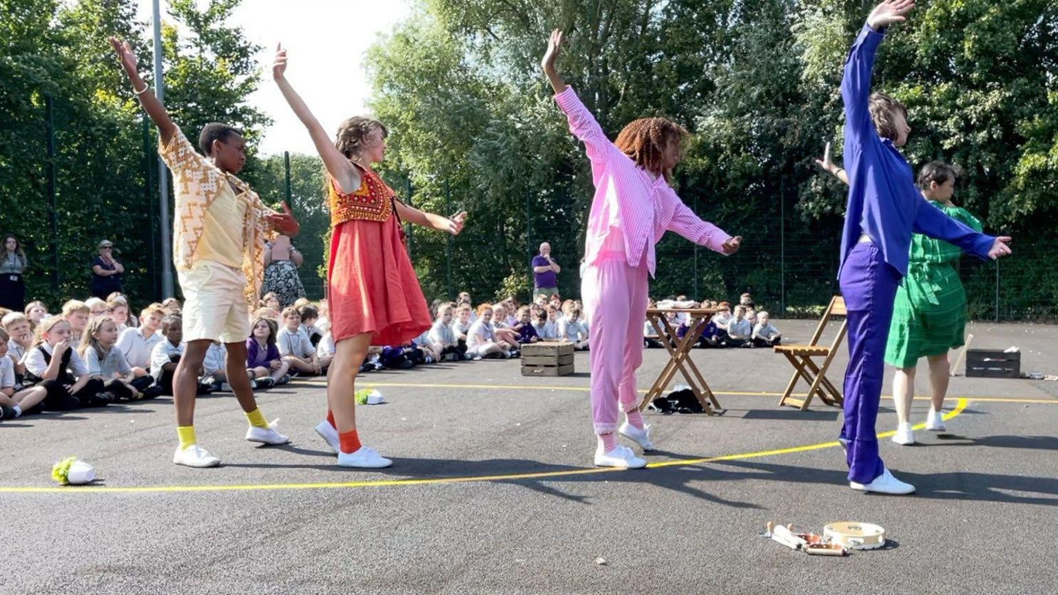 Five dancers dressed in colourful clothes (cream, red, pink, blue, green) doing ballet on a school playground with children sitting watching in the background