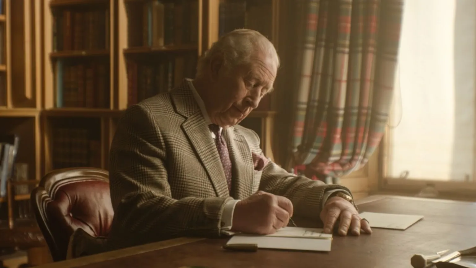 King Charles III writes a letter from a desk in front of a window in Buckingham Palace. He is wearing a blazer and a purple tie.
