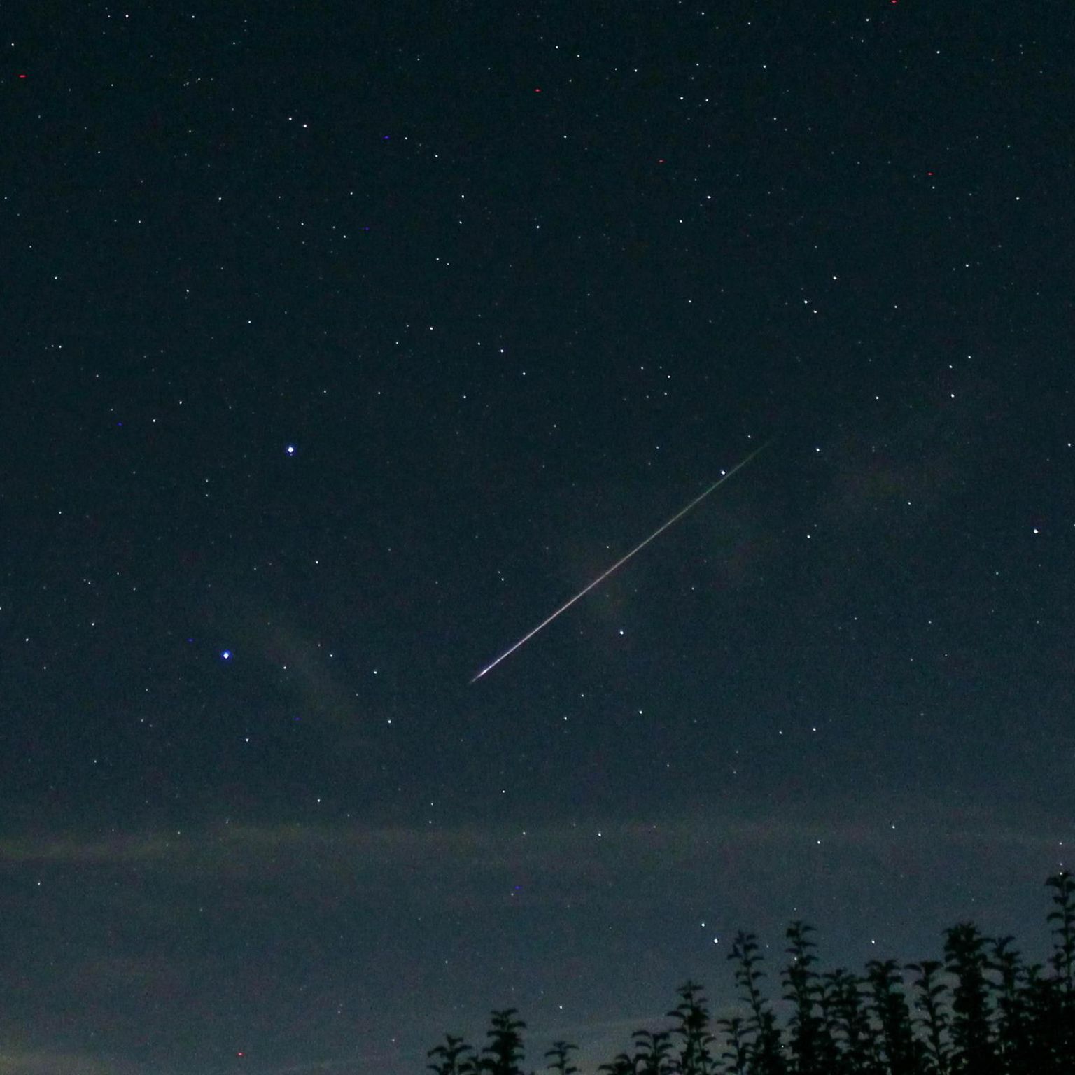 A long white line streaks across a dark blue sky which is dotted with stars