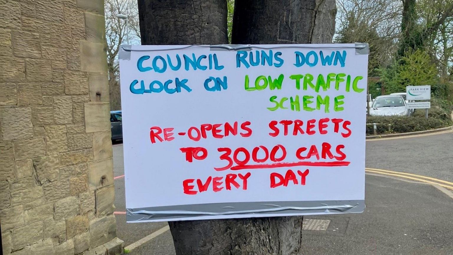Placard on tree reading 'council runs down clock on low traffic scheme' and 're-opens streets to 3000 cars every day'