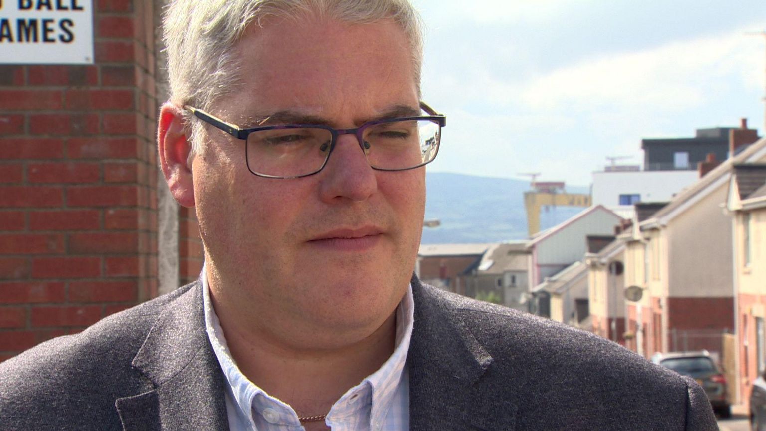 Gavin Robinson standing in a street. Houses are in the background as is one of the H&W cranes. Mr Robinson is wearing a checked blue and white shirt, a grey blazer and glasses.