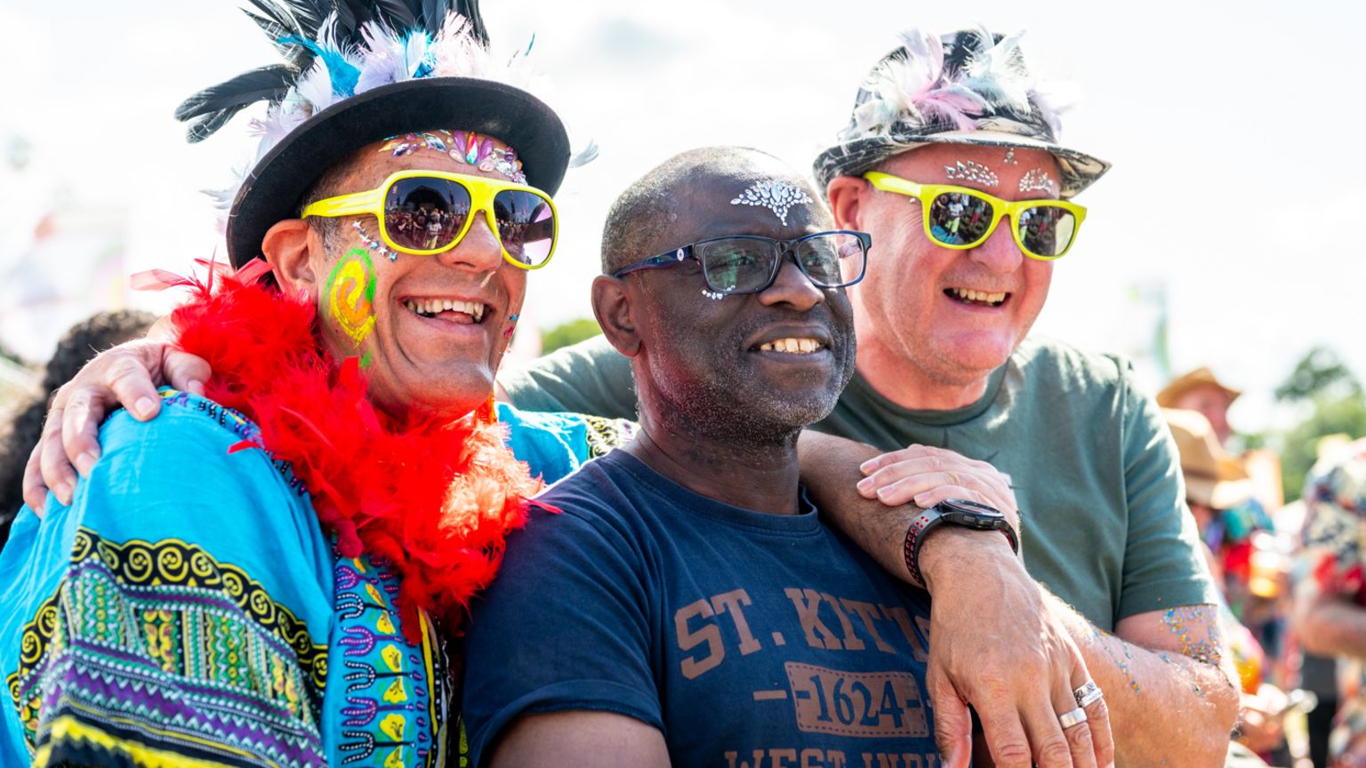 Three people wearing glasses and yellow sunglasses with diamantes and stubble on their painted faces smiling