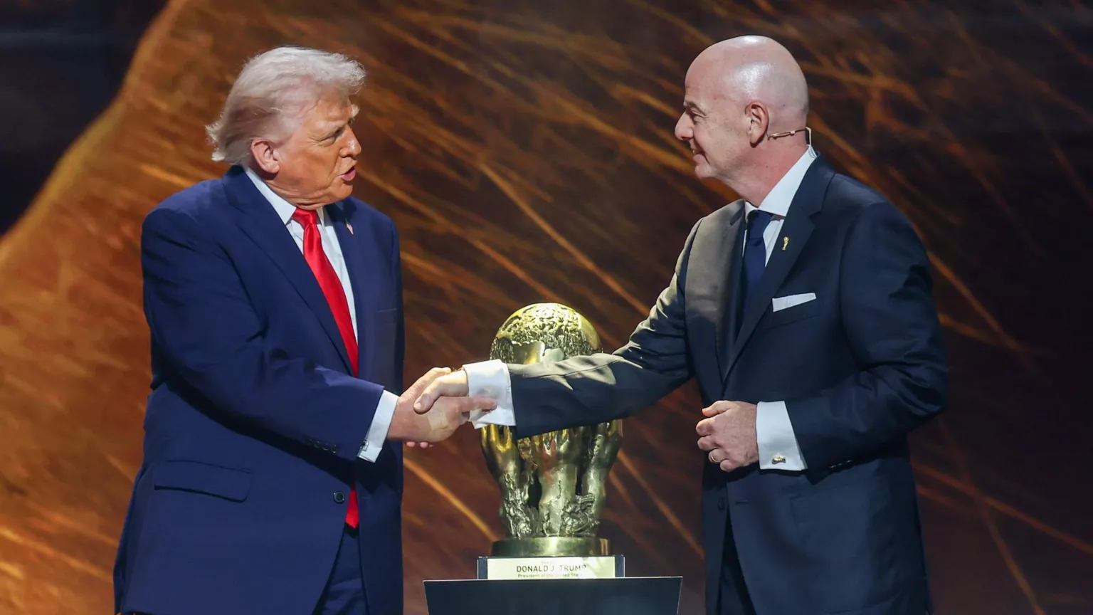 Getty Images US President Donald Trump, wearing a navy blue suit, white shirt and red tie, shakes hands with Gianni Infantino, wearing a dark blue suit, white shirt and tie as both men stand on stage in front of the Fifa Peace Prize which is on a plinth