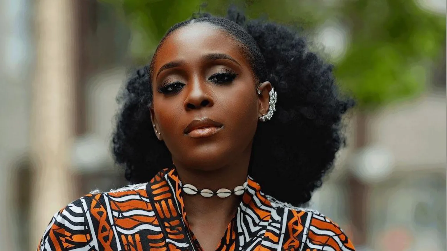 Supplied Portrait of a woman standing outdoors in a black, white, and orange patterned garment with a shell necklace and decorative ear cuff. Trees and buildings are softly blurred in the background.